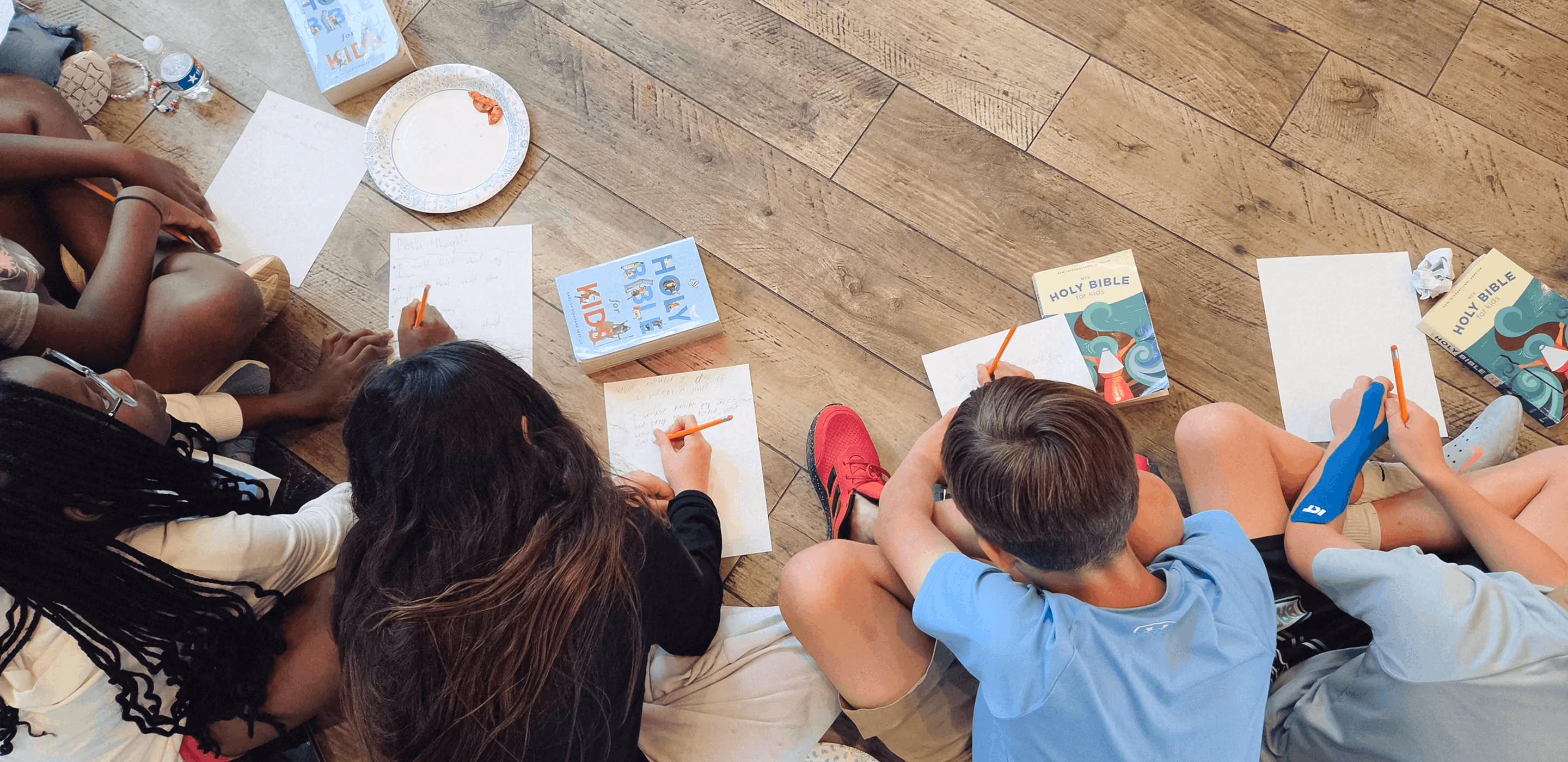 Children sitting on floor writing on paper with pencils, surrounded by 'Holy Bible for Kids' books.