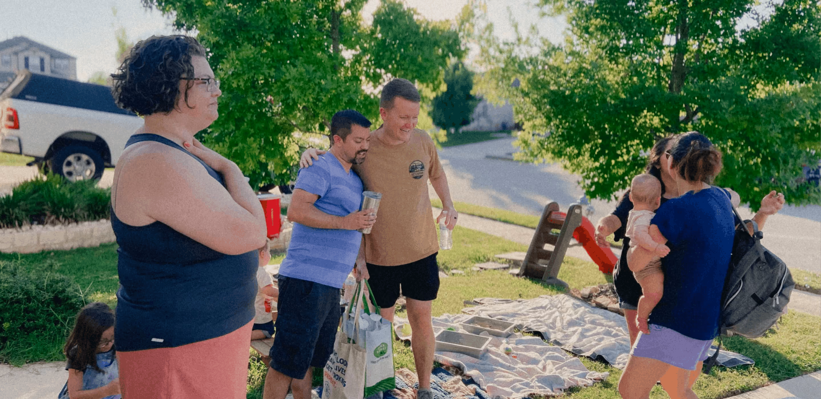 Group of adults and children enjoying an outdoor gathering with grass and trees in a neighborhood.