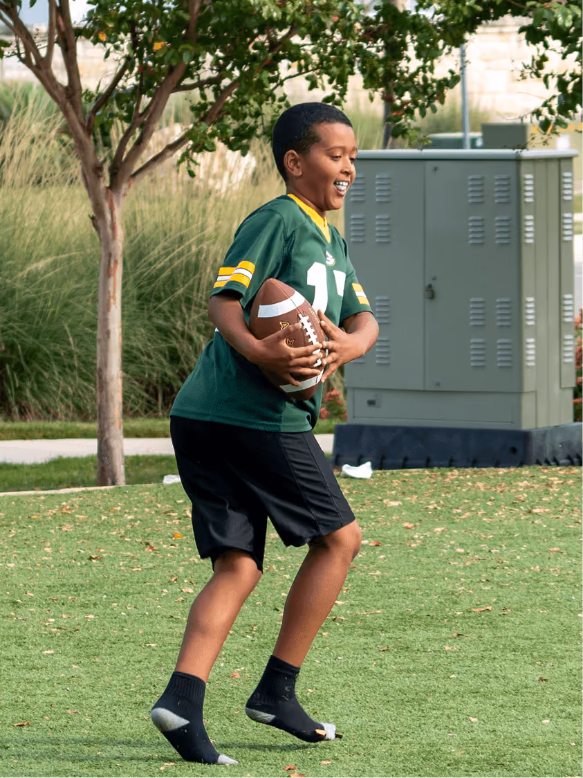 Boy smiling while running barefoot on grass holding a football near trees and electrical box.