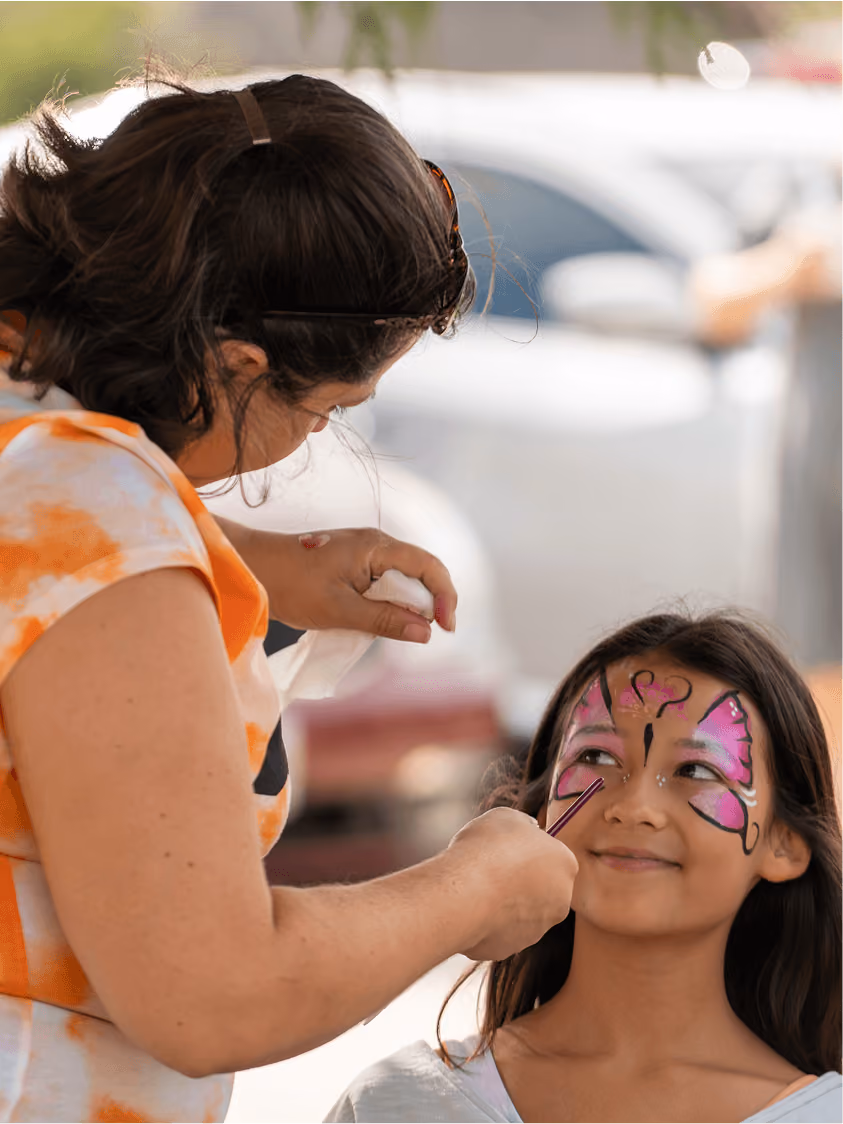 Woman painting pink butterfly face art on a smiling girl's face outdoors.