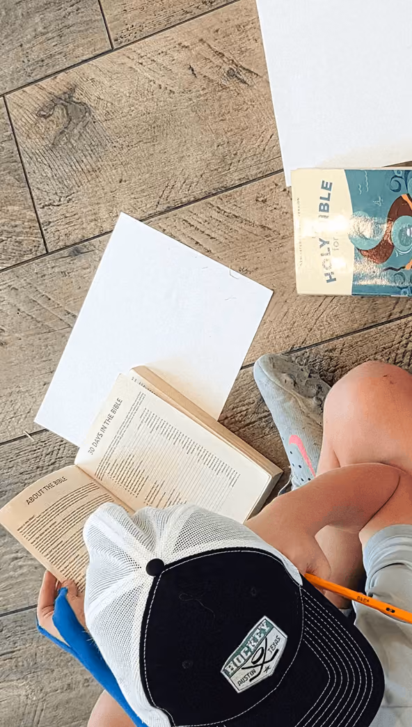 Overhead view of a child reading an open Bible with blank papers on a wood floor.