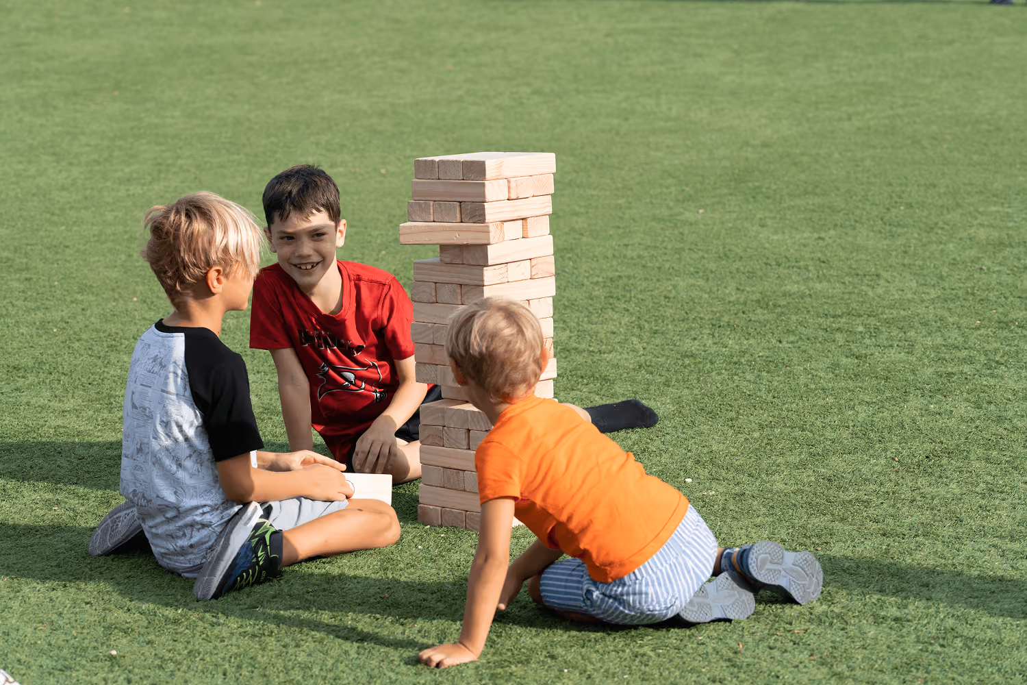 Three boys sitting on grass playing a large wooden block stacking game outdoors.