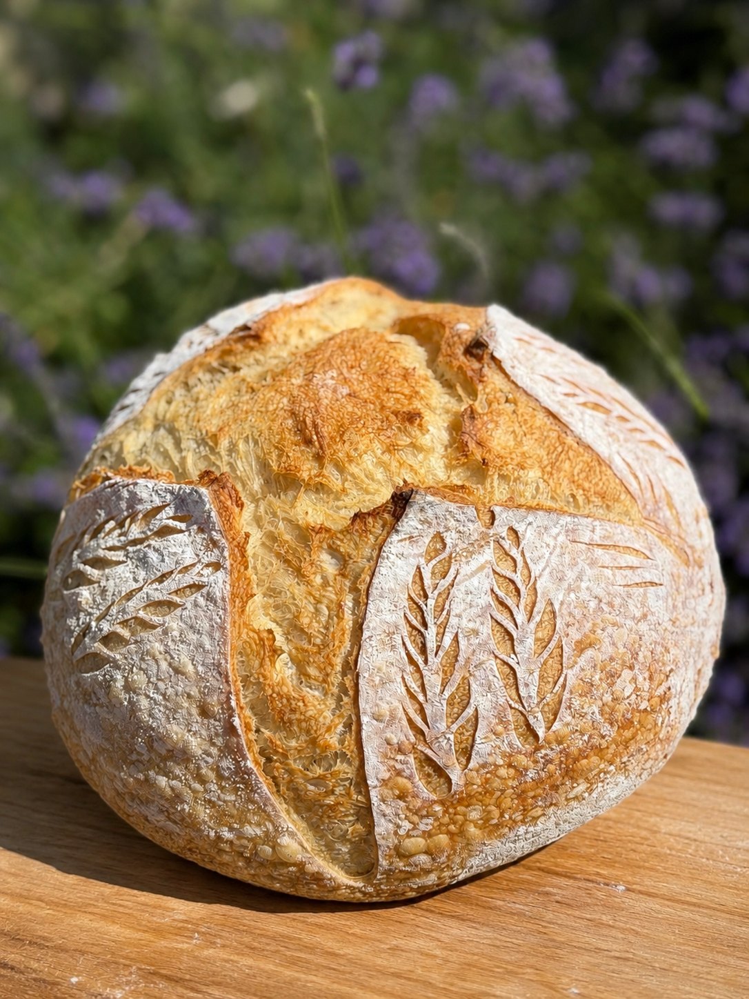 Round sourdough loaf scored with wheat stalks, cooling on a wood board beside blooming lavender