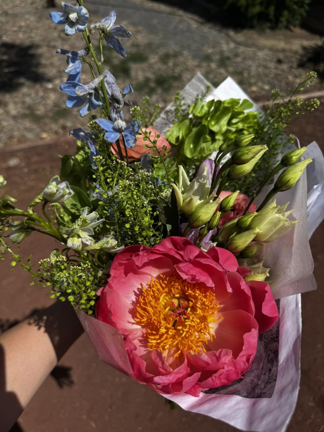 A wrapped farm bouquet with a coral peony, blue delphinium and bells of Ireland