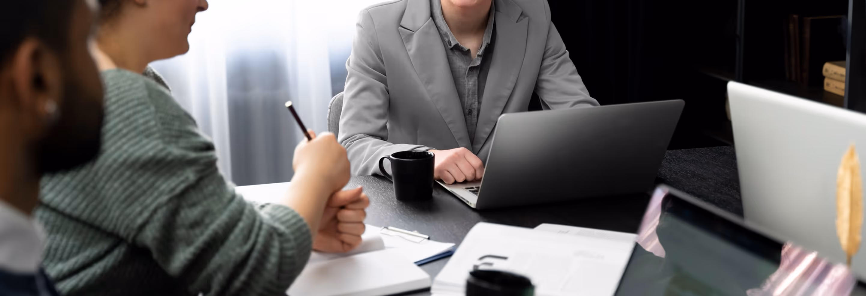 Three people in a meeting around a table with laptops, notebooks, and coffee mugs.
