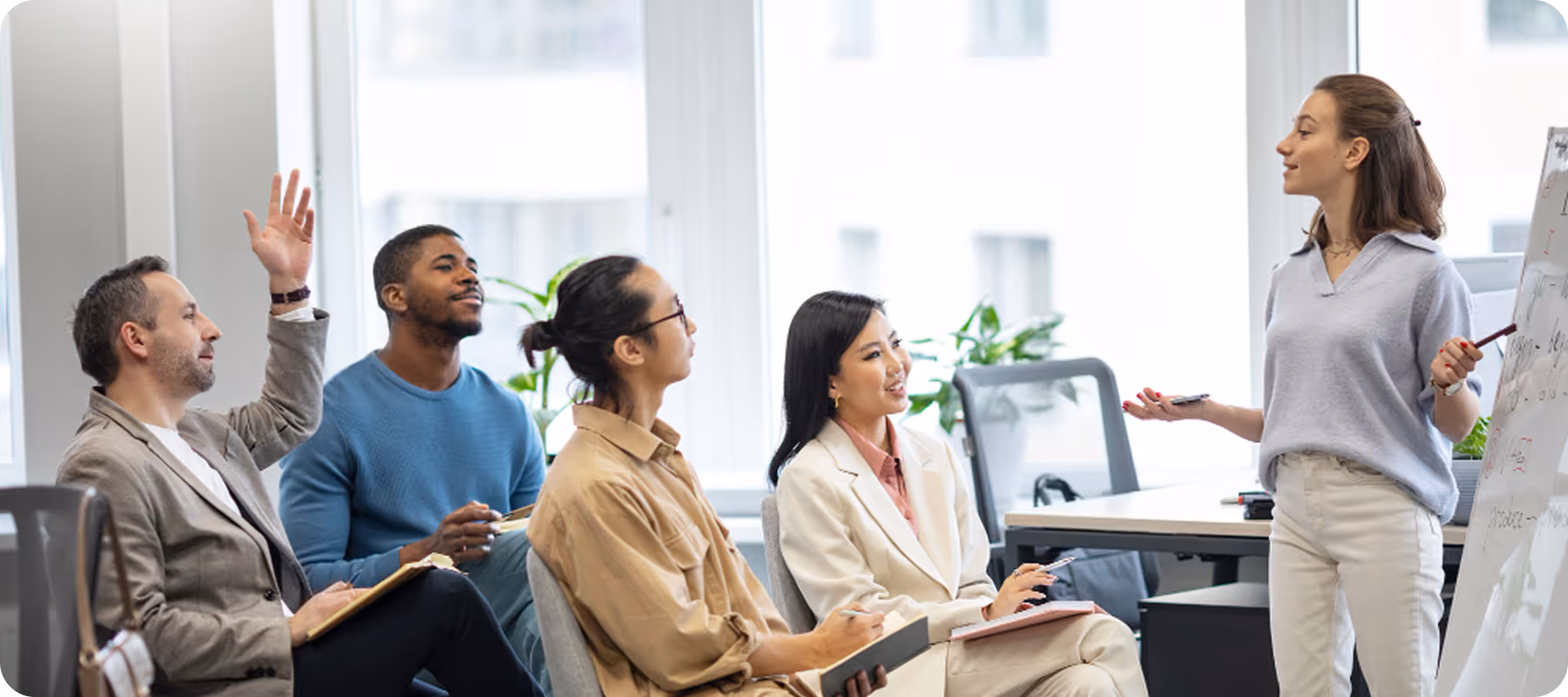 A woman presents ideas on a whiteboard to a group of attentive colleagues seated in an office.