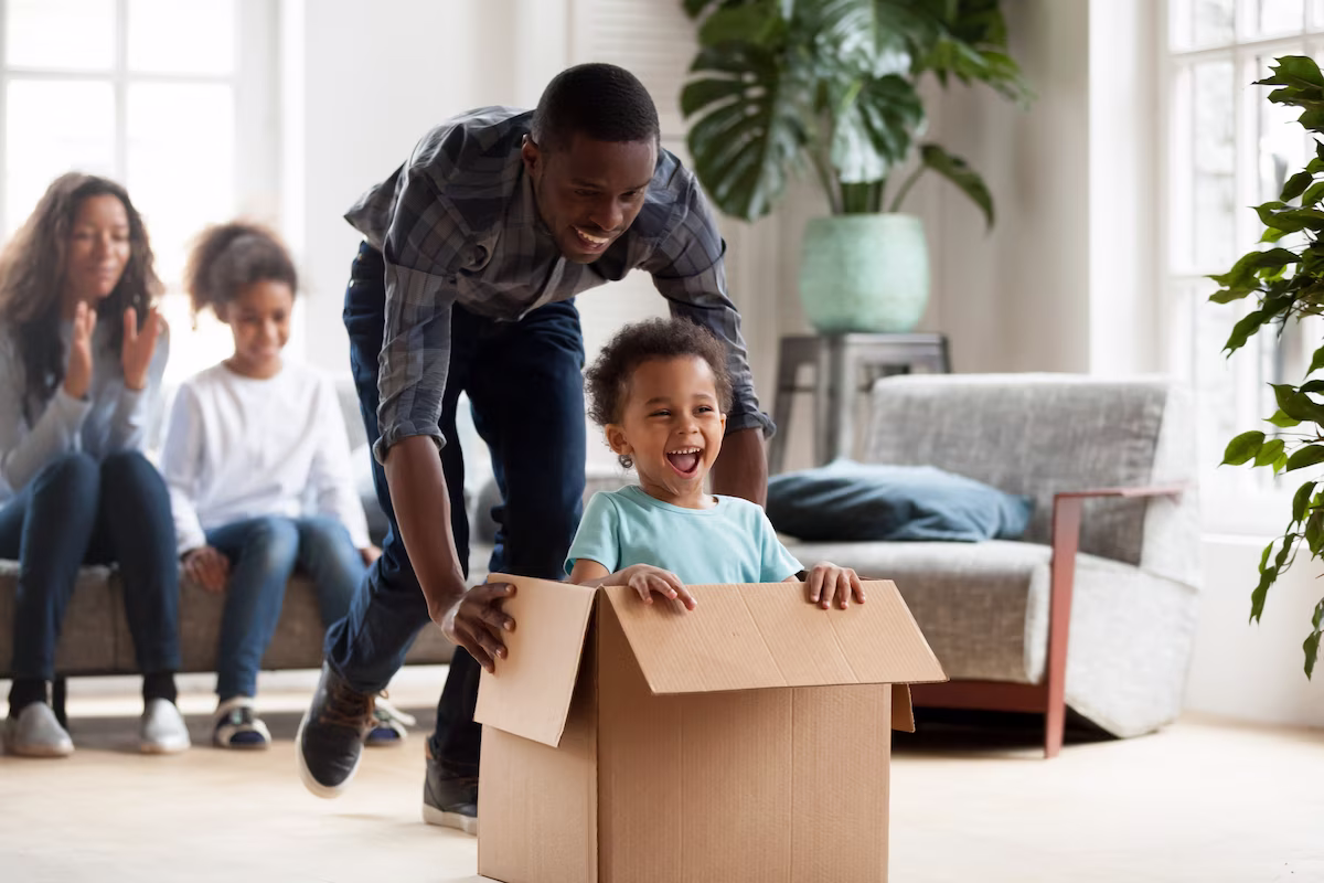 A father plays with his son in a cardboard box while the family cheers