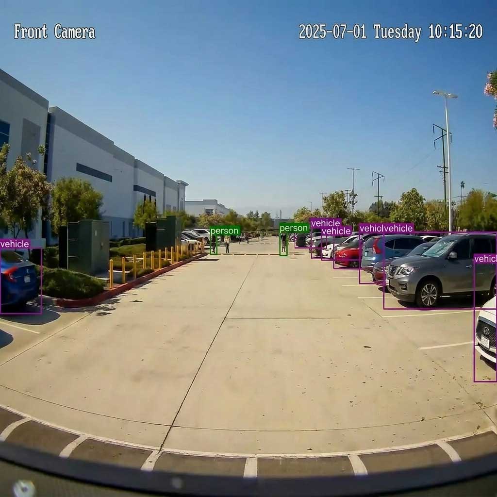 Security camera footage of a person walking on a paved lot at night near blue fencing and parked vehicles.
