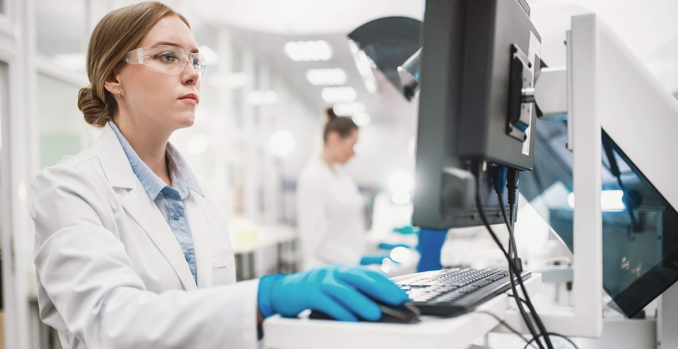 Scientist wearing safety glasses and gloves working on a computer in a bright laboratory.