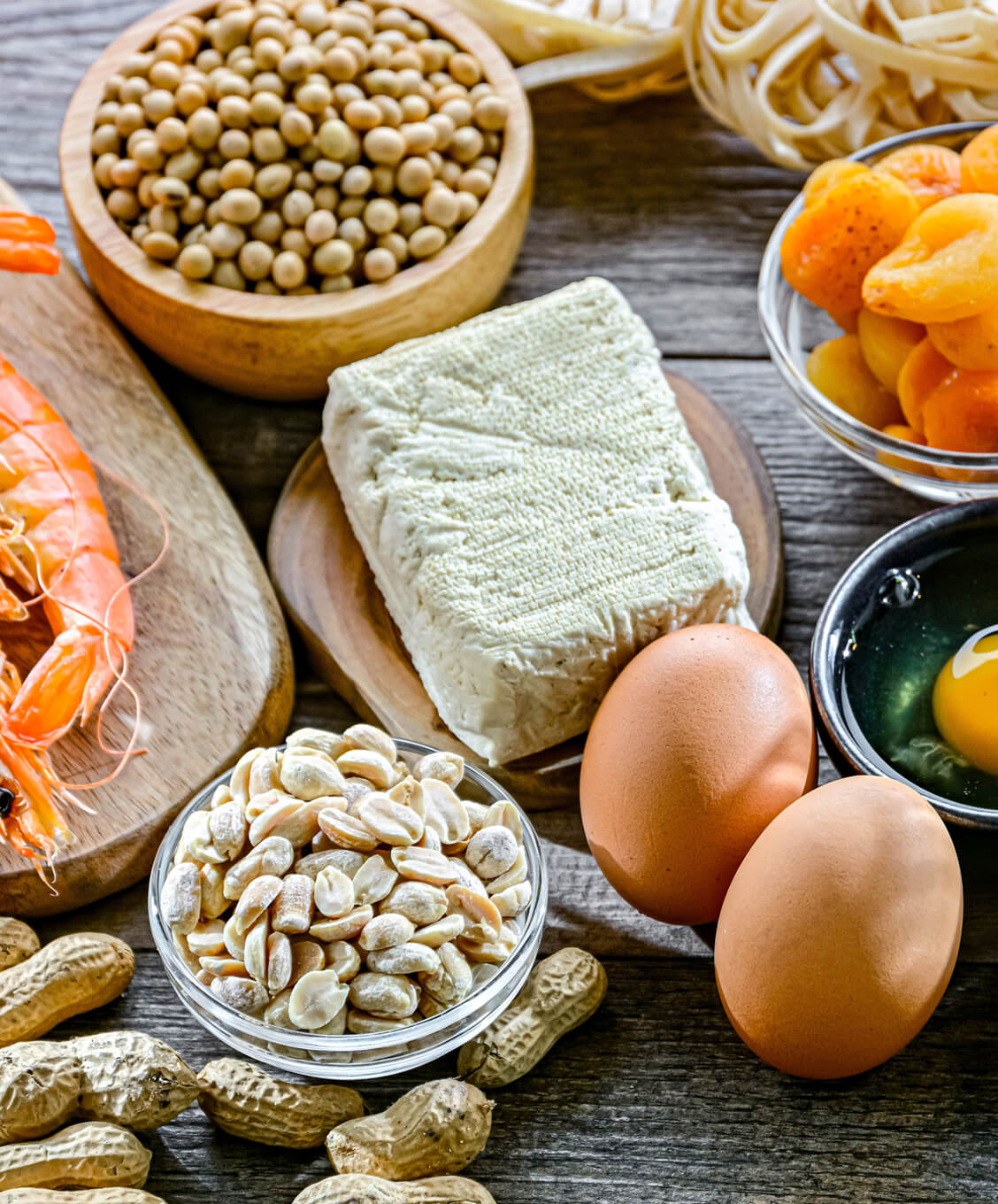 Variety of allergenic foods including shrimp, soybeans, tofu, peanuts, eggs, pasta, and dried apricots on wood table.
