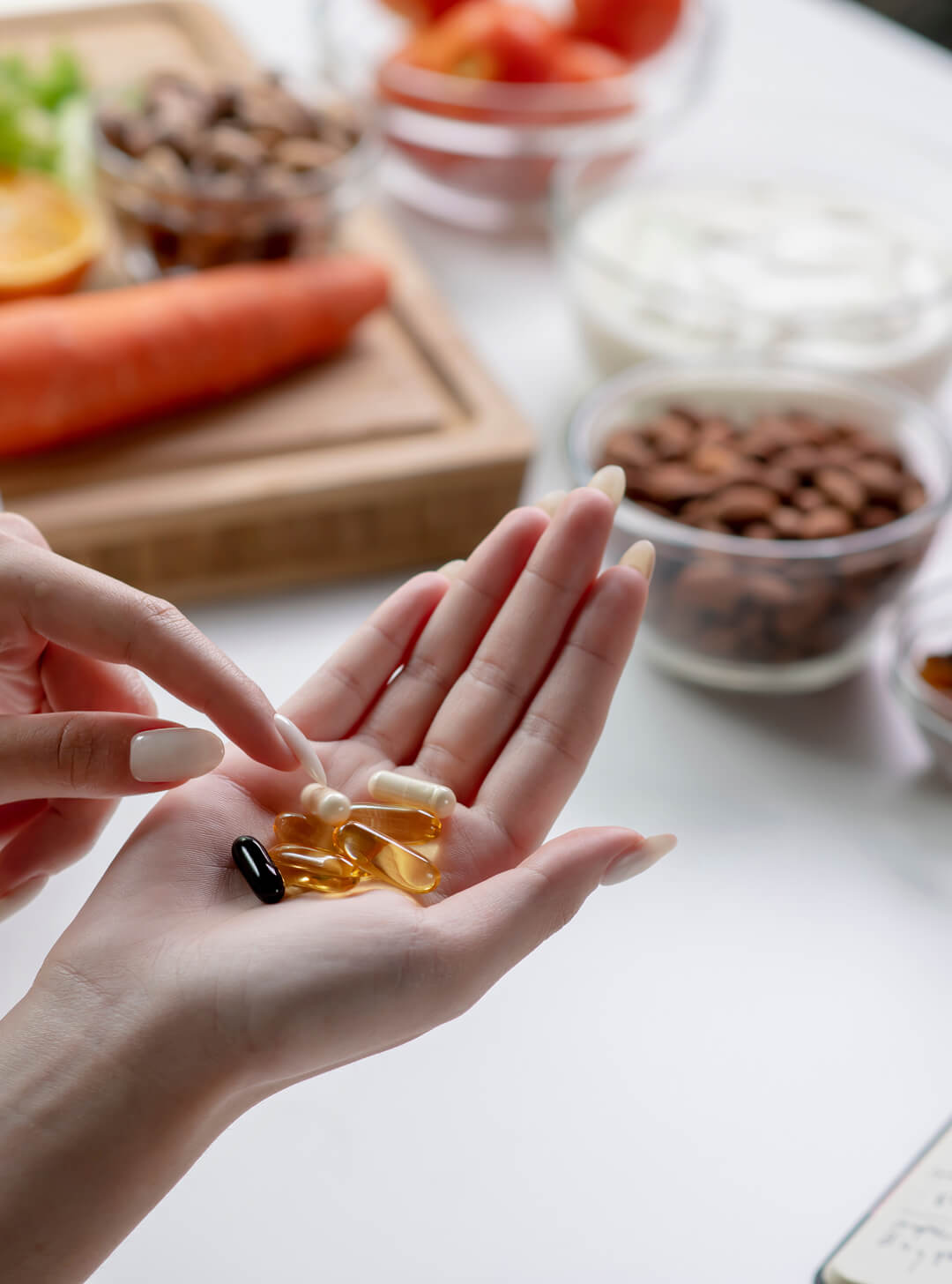 Hand holding several capsules and softgel supplements with a blurred background of nuts and vegetables.