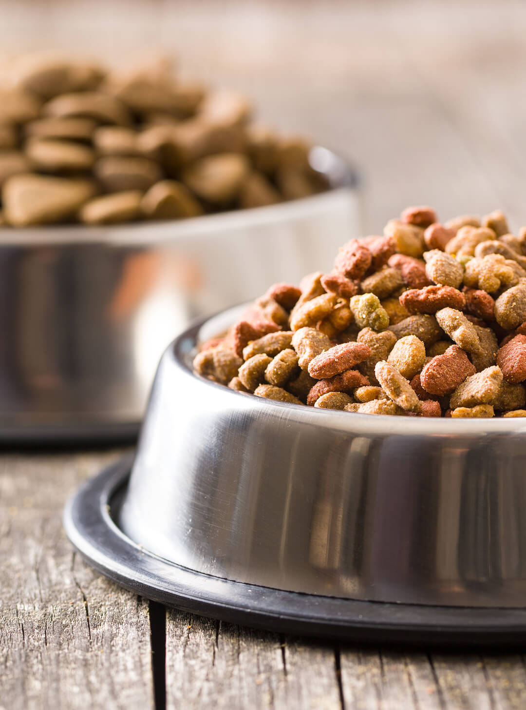 Two metal bowls filled with dry pet food on a wooden surface, one with multicolored kibble.