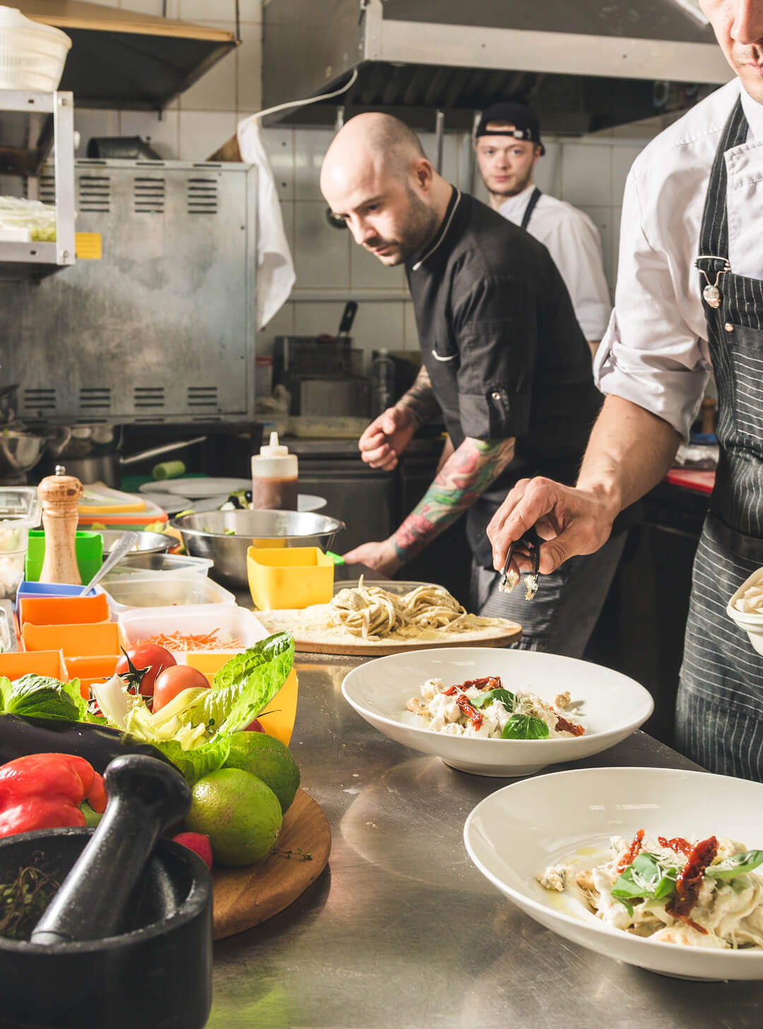 Chefs preparing and plating pasta dishes in a busy commercial kitchen with fresh vegetables nearby.