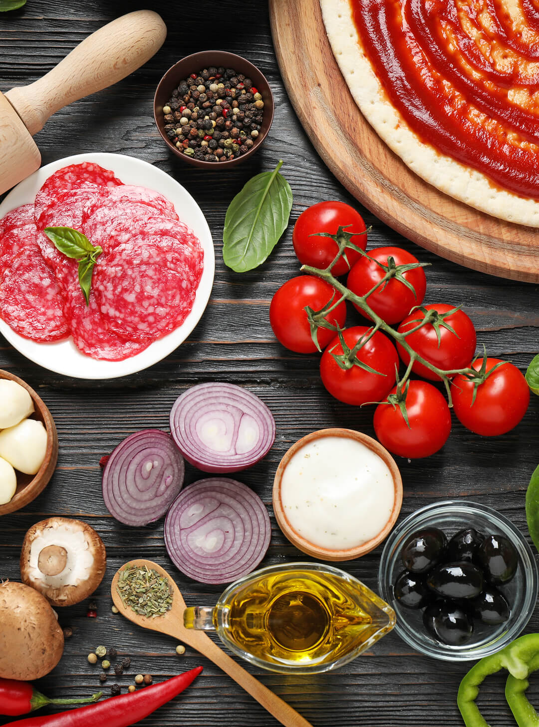Pizza dough with tomato sauce, cherry tomatoes, salami, onions, olives, garlic, mushrooms, oil, herbs, and basil leaves.