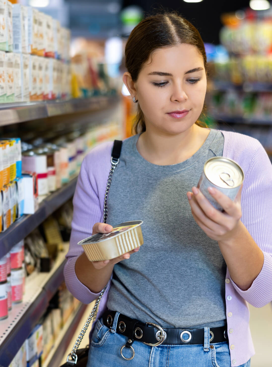 Woman in a lilac cardigan compares two canned food items in a grocery store aisle.