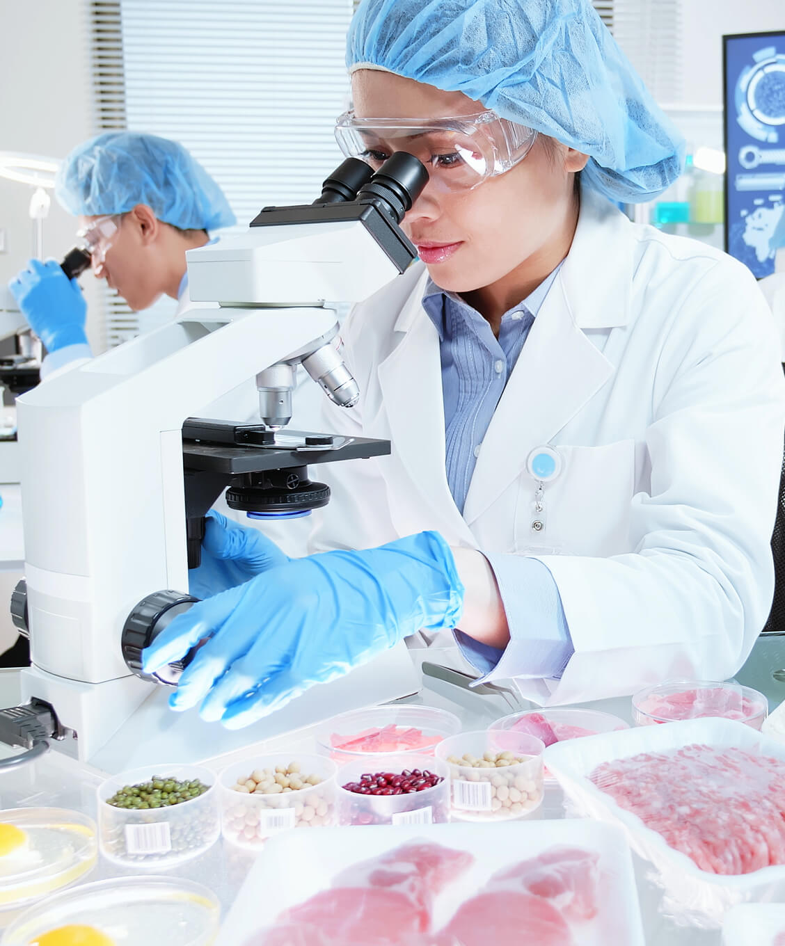 Scientists in lab coats and gloves examining samples under microscopes with packaged meats nearby.
