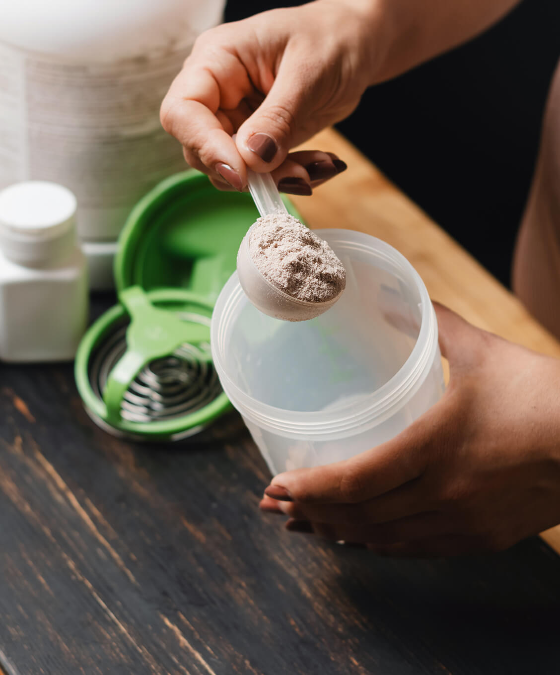 Person scooping protein powder into a clear shaker bottle with supplement containers nearby.