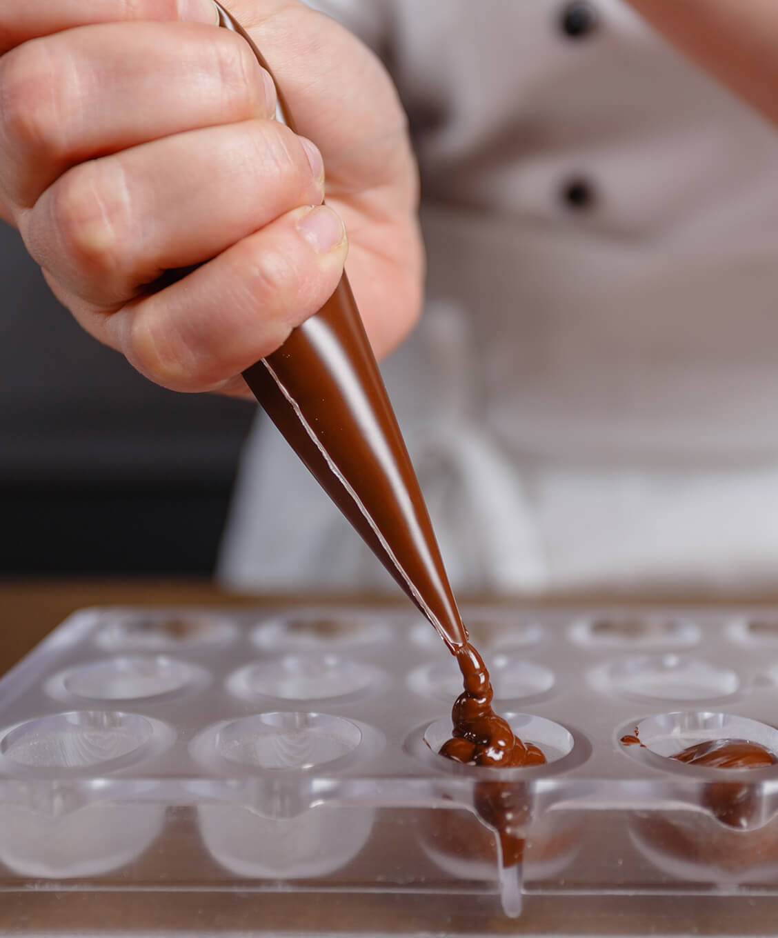 Hand with piping bag filling a plastic mold with melted chocolate for making chocolates.