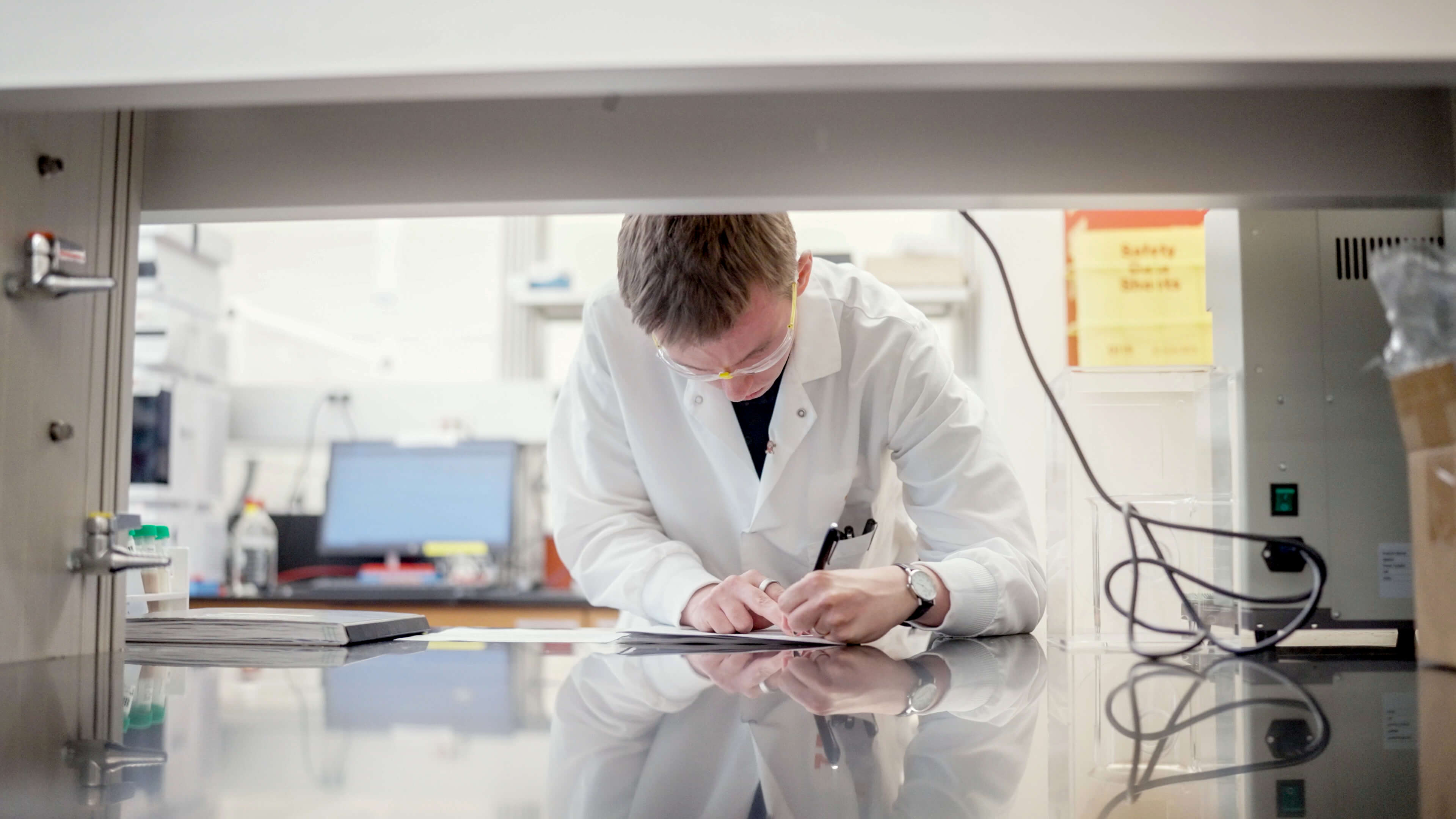 Scientist wearing a white lab coat and safety glasses writing notes on paper at a reflective laboratory bench.
