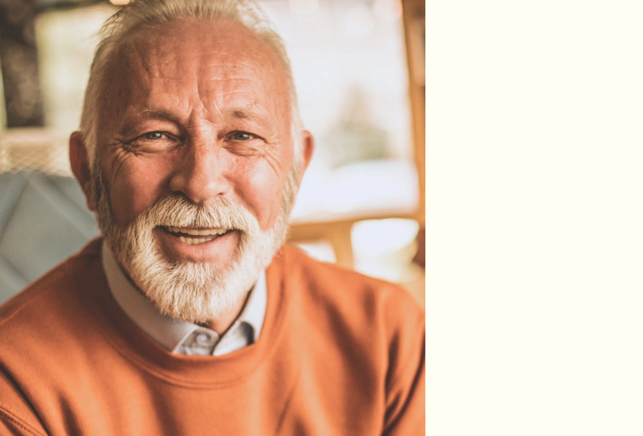 Older man with white beard wearing orange t-shirt smiling at camera with blurred interior background and cream-colored right side