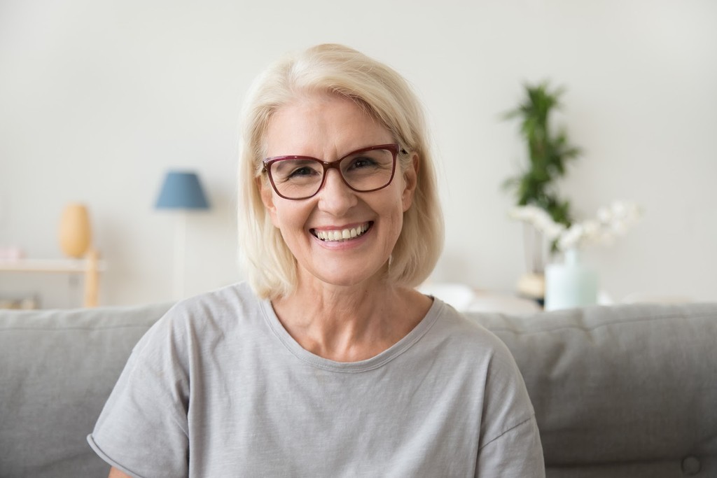 Smiling middle aged mature grey haired woman looking at camera, sitting on sofa in living room headshot portrait