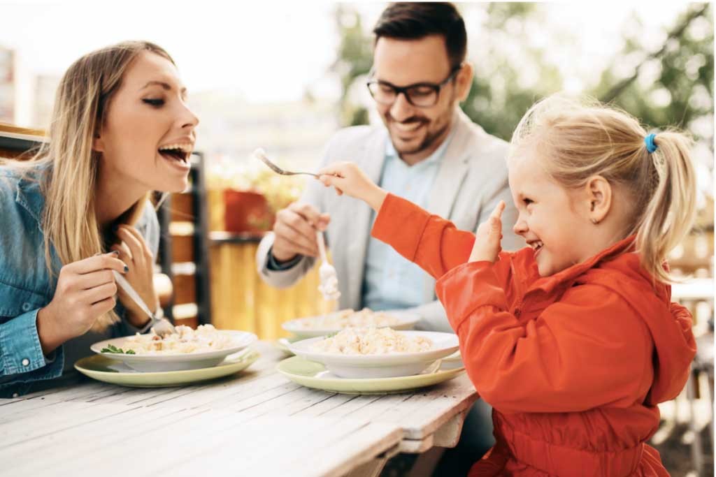 Family of three happily eating together at an outdoor table on a sunny day