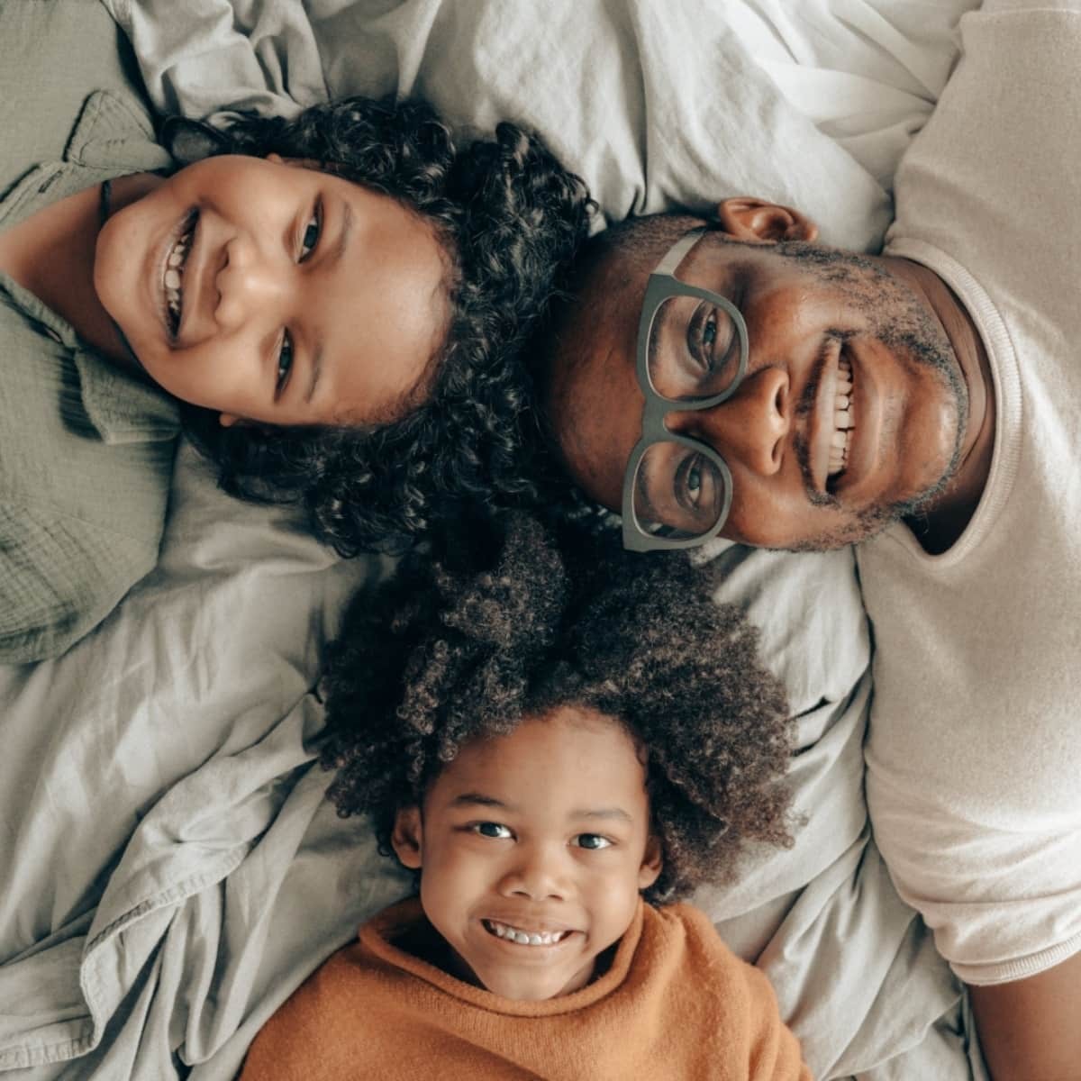 Family of three smiling together lying on white bedding, viewed from above