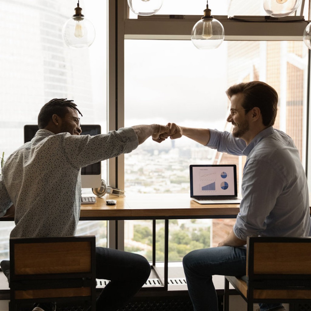 Two colleagues fist bumping at desk with laptop and city view through office windows