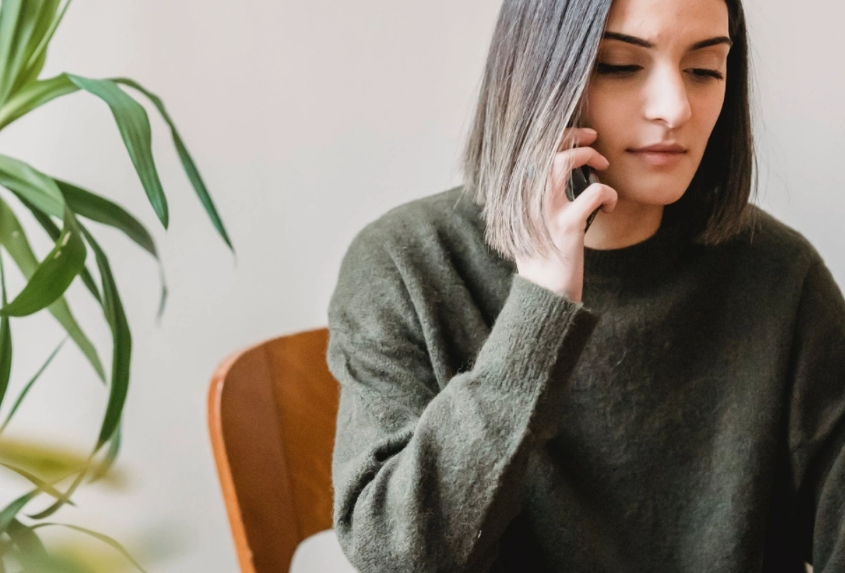 Woman in olive sweater sitting on orange chair with hand to face, green plant beside her