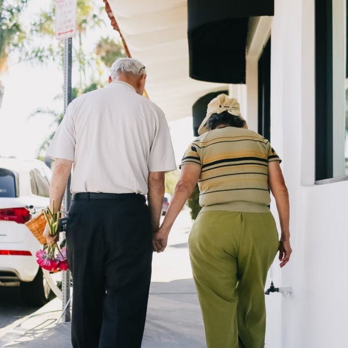 Older man and person holding flowers walking together toward white building with modern architecture