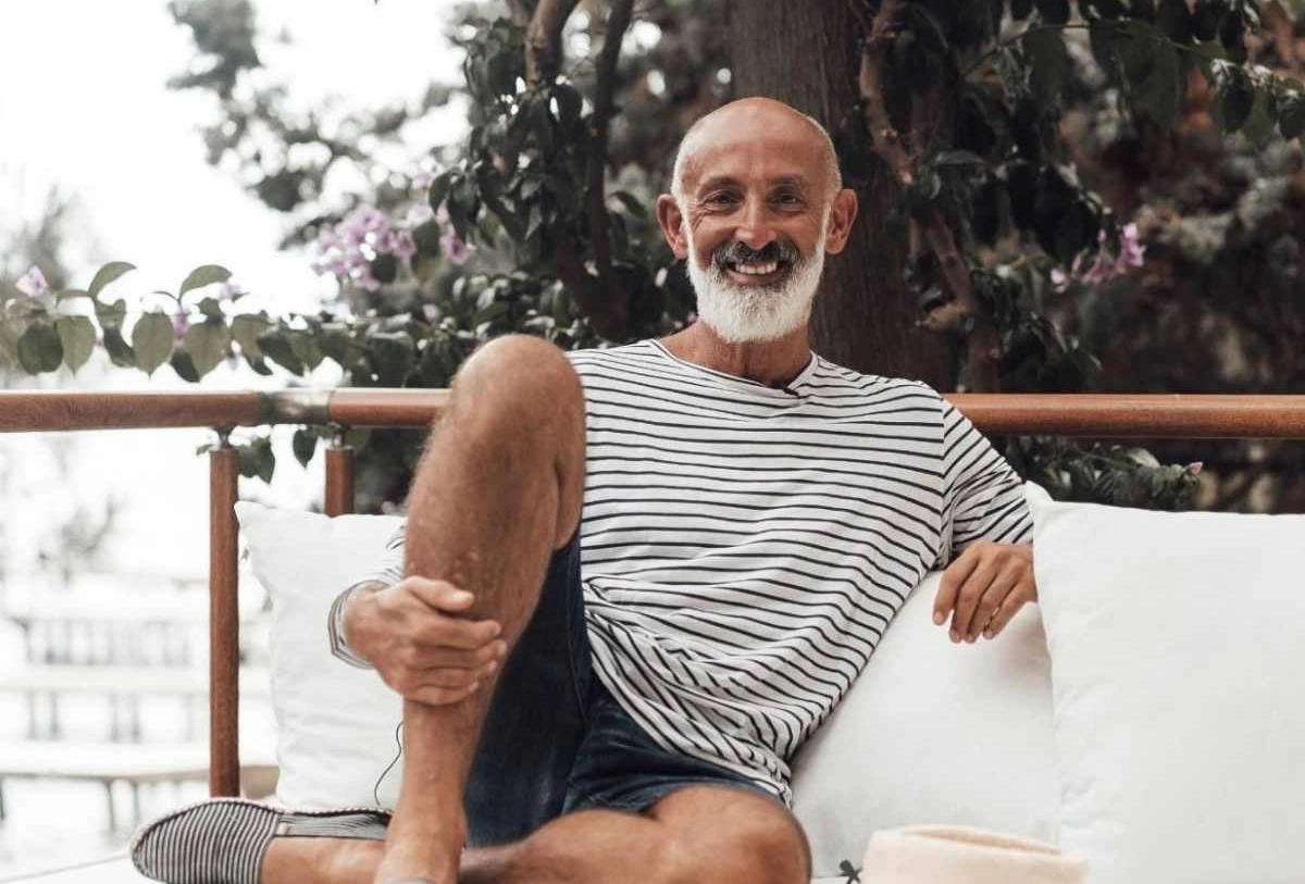 Smiling older man with white beard wearing striped shirt sitting outdoors with flowering plants