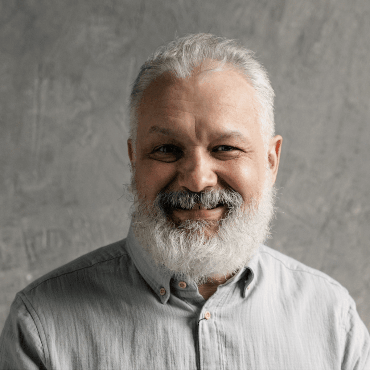 Smiling older man with gray beard wearing button-up shirt against gray textured backdrop