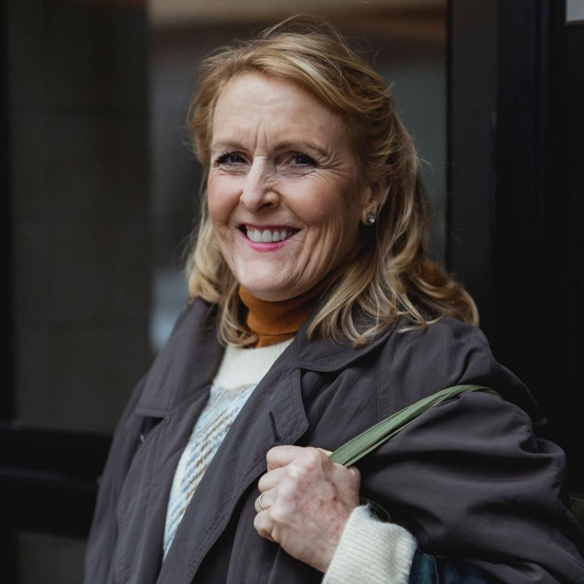 Woman in black blazer over brown turtleneck holding green folder, smiling indoors