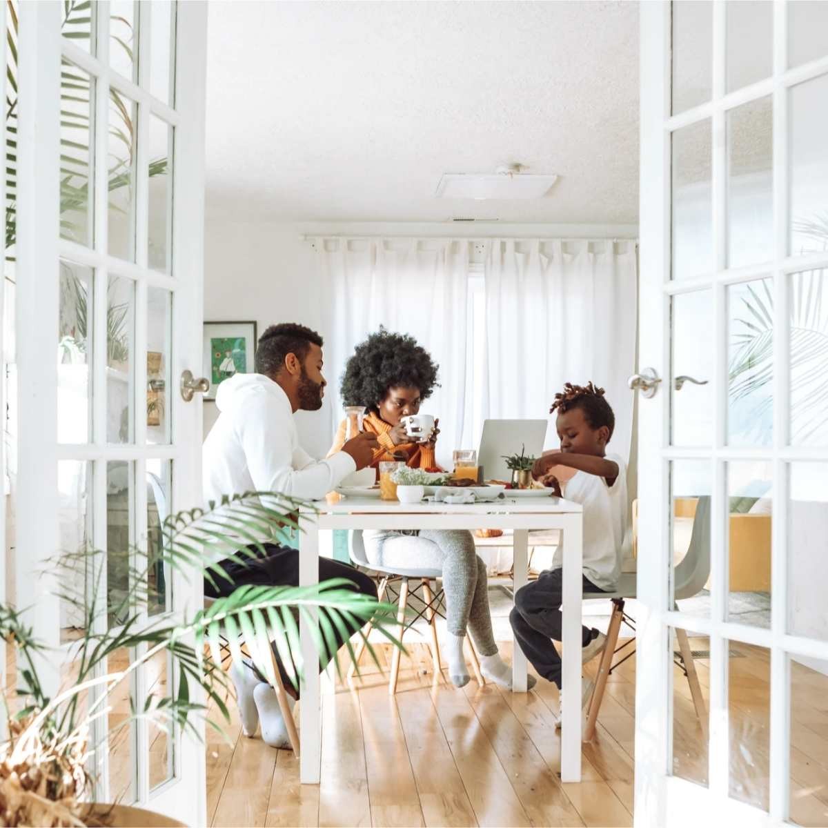 Family dining together at white table in bright room with plants and glass doors, one child using laptop