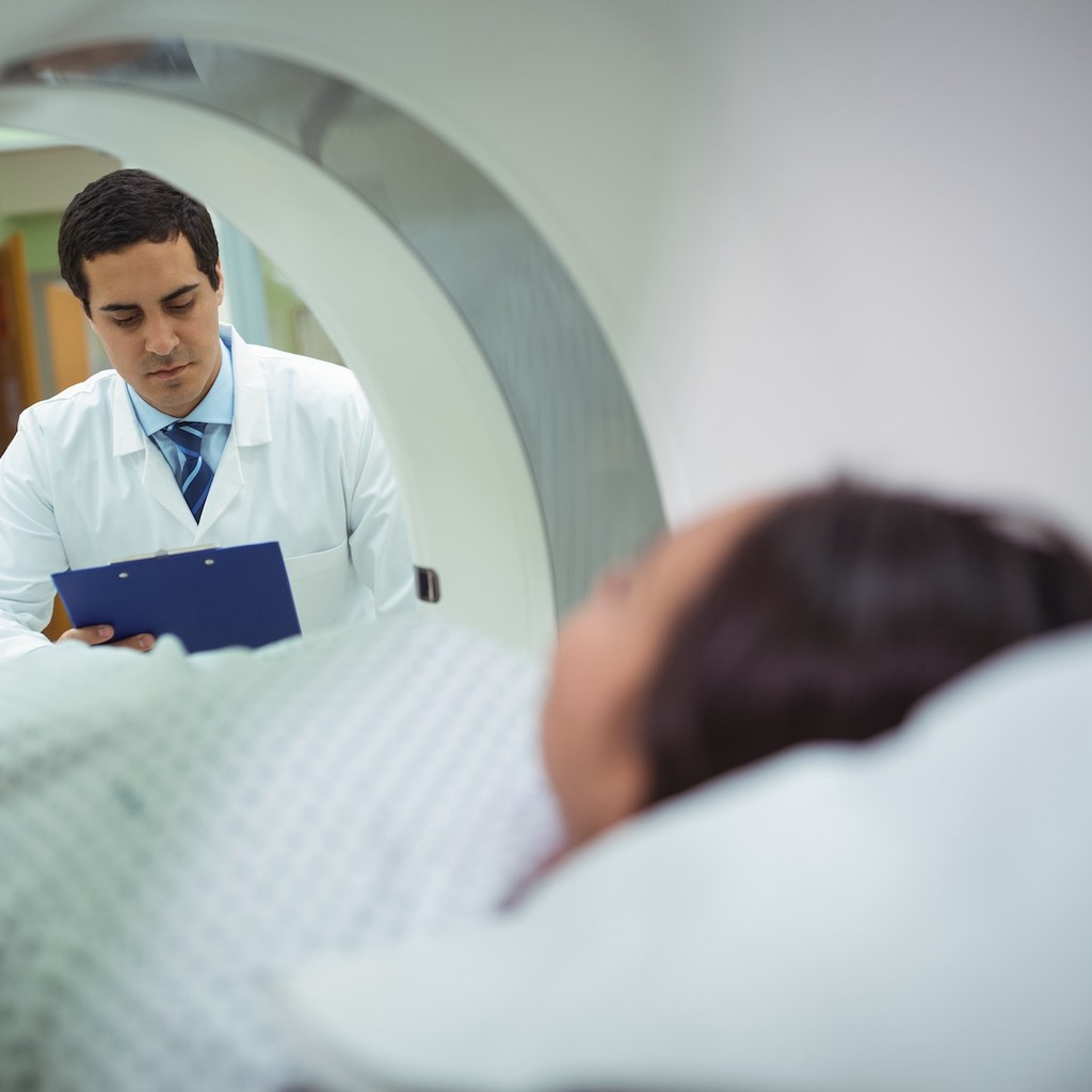 Doctor in white coat reviewing clipboard while patient positioned in CT or MRI scanner for diagnostic imaging