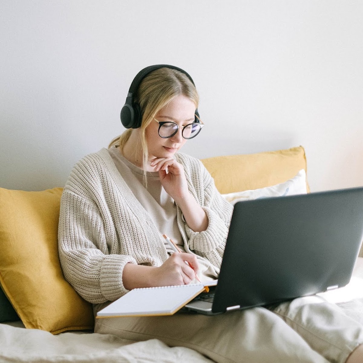 Woman wearing glasses and headphones working on laptop while seated on yellow chair with notebook