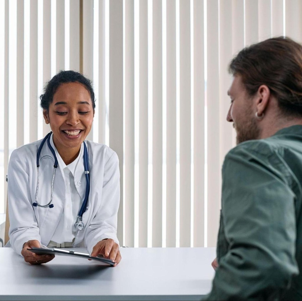 Female doctor in white coat with stethoscope smiling while consulting with male patient at desk