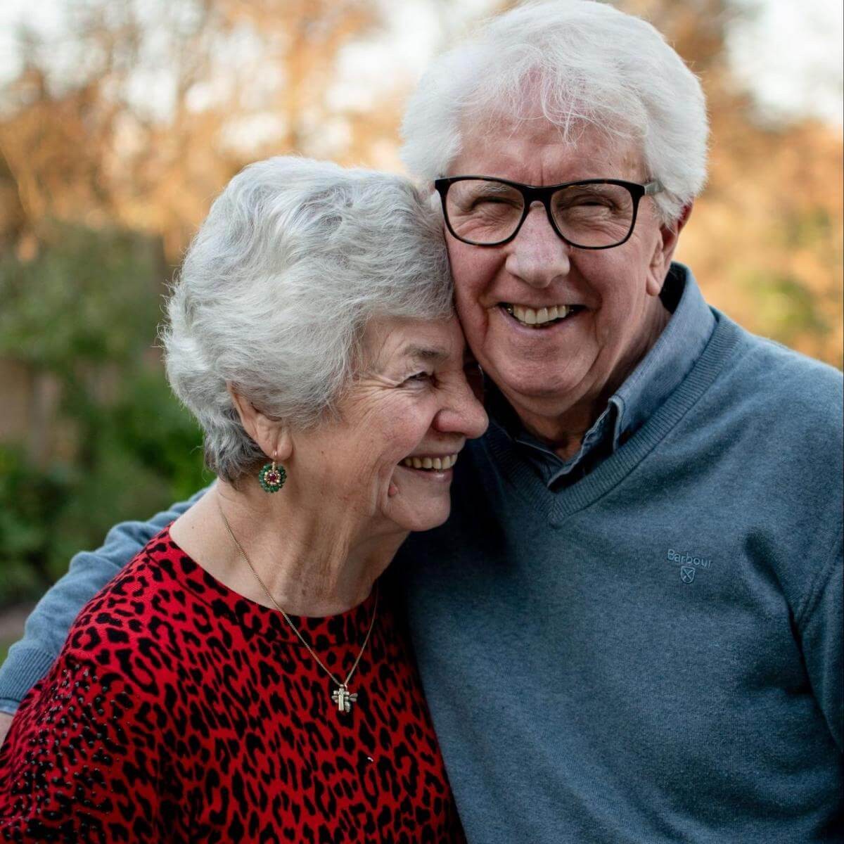 Smiling senior couple embracing outdoors with autumn background, woman in red patterned top and man in blue sweater