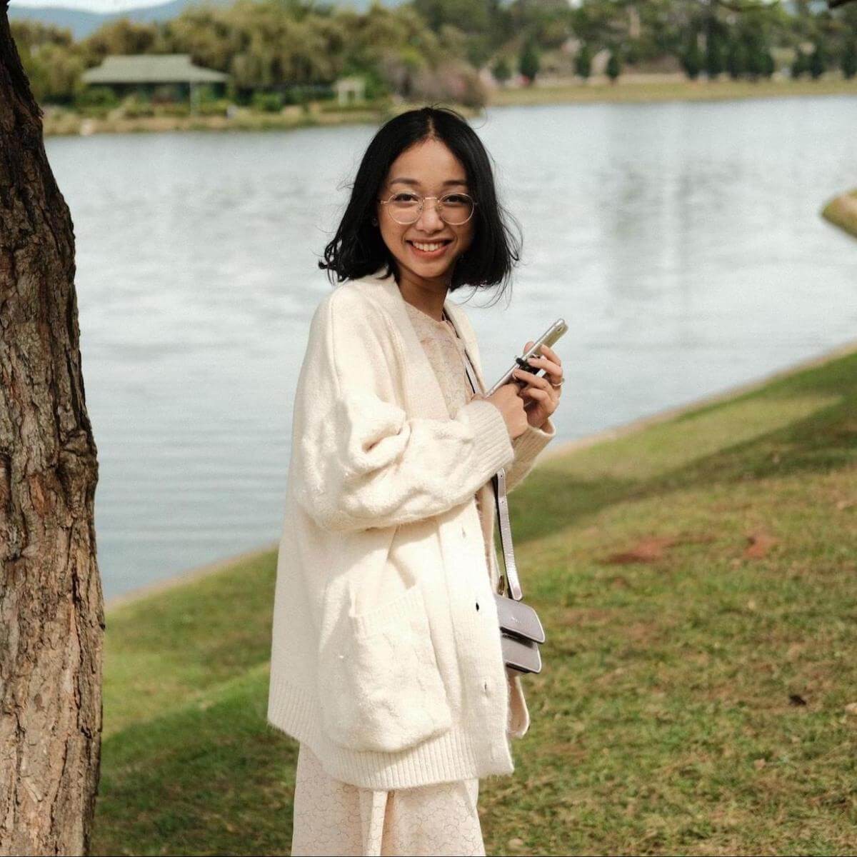 Woman in cream cardigan holding phone while standing by tree overlooking calm lake and forest