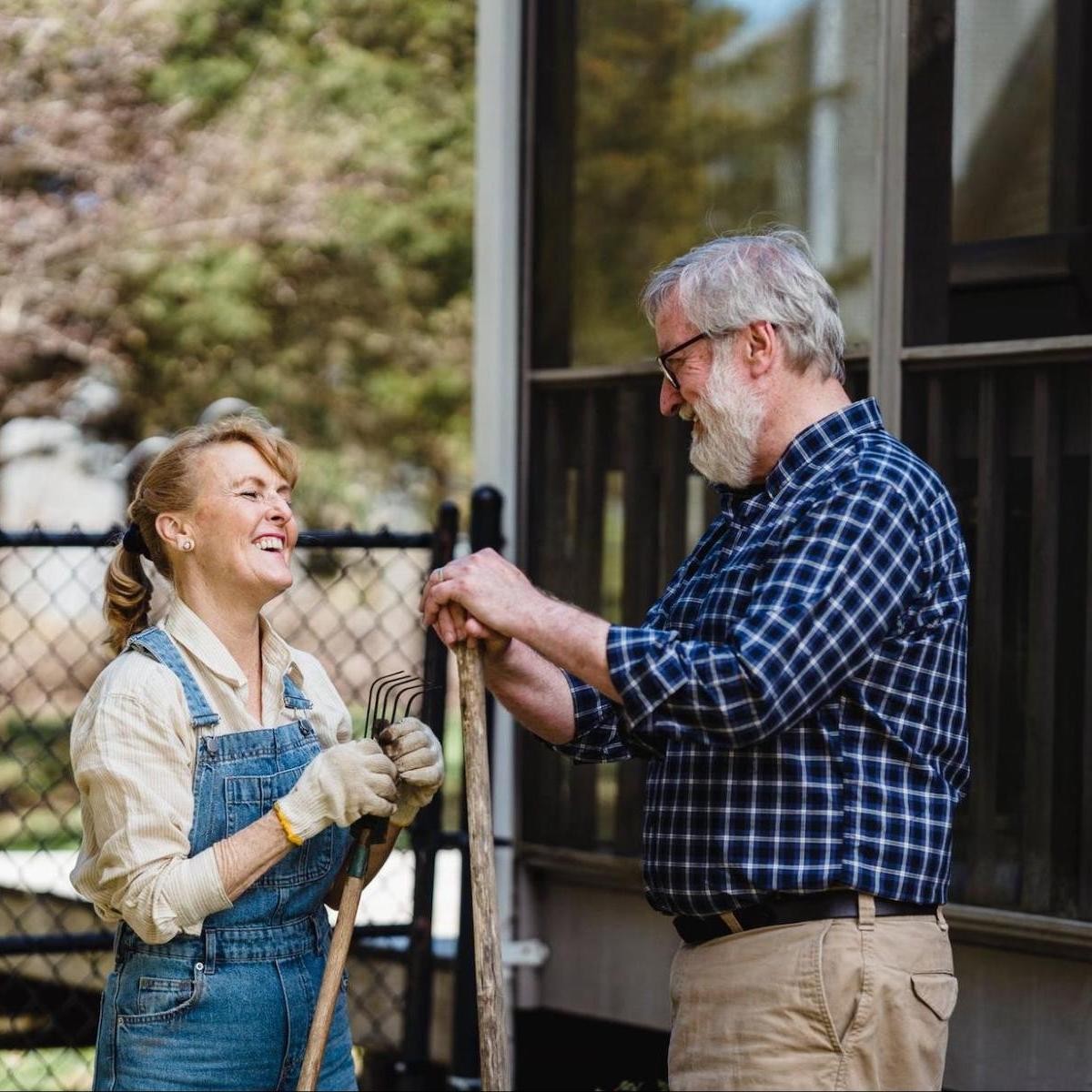 Older couple smiling while gardening, woman in denim overalls and man in blue plaid shirt holding rakes outside house