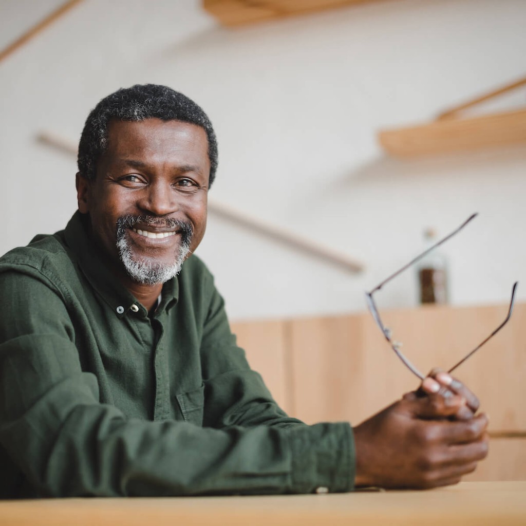 Smiling man in green shirt holding glasses indoors with wooden background