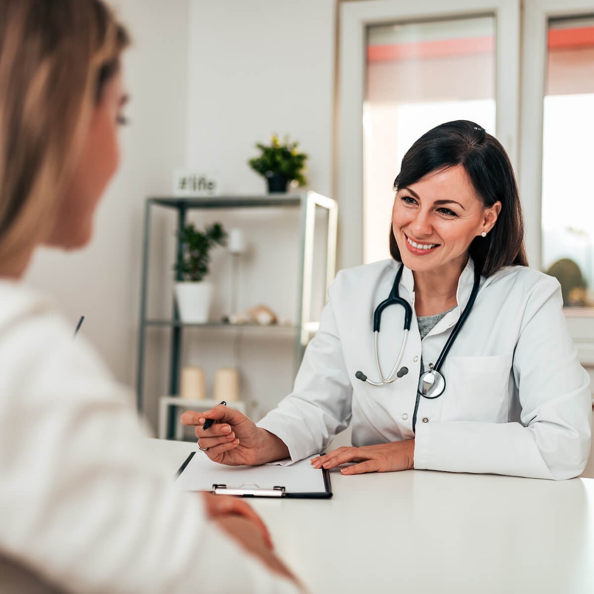Female doctor in white coat with stethoscope smiling at patient during consultation at desk in modern office