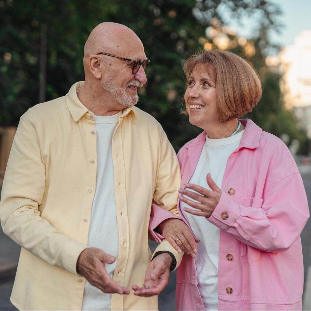 Senior couple smiling and walking together outdoors, man in yellow shirt and woman in pink jacket with trees in background