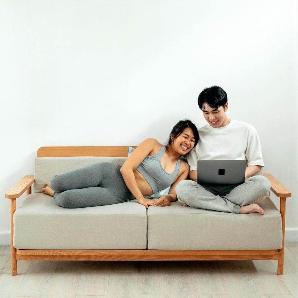 Couple on beige couch with wooden frame, woman in gray athletic wear, man in white shirt viewing laptop together