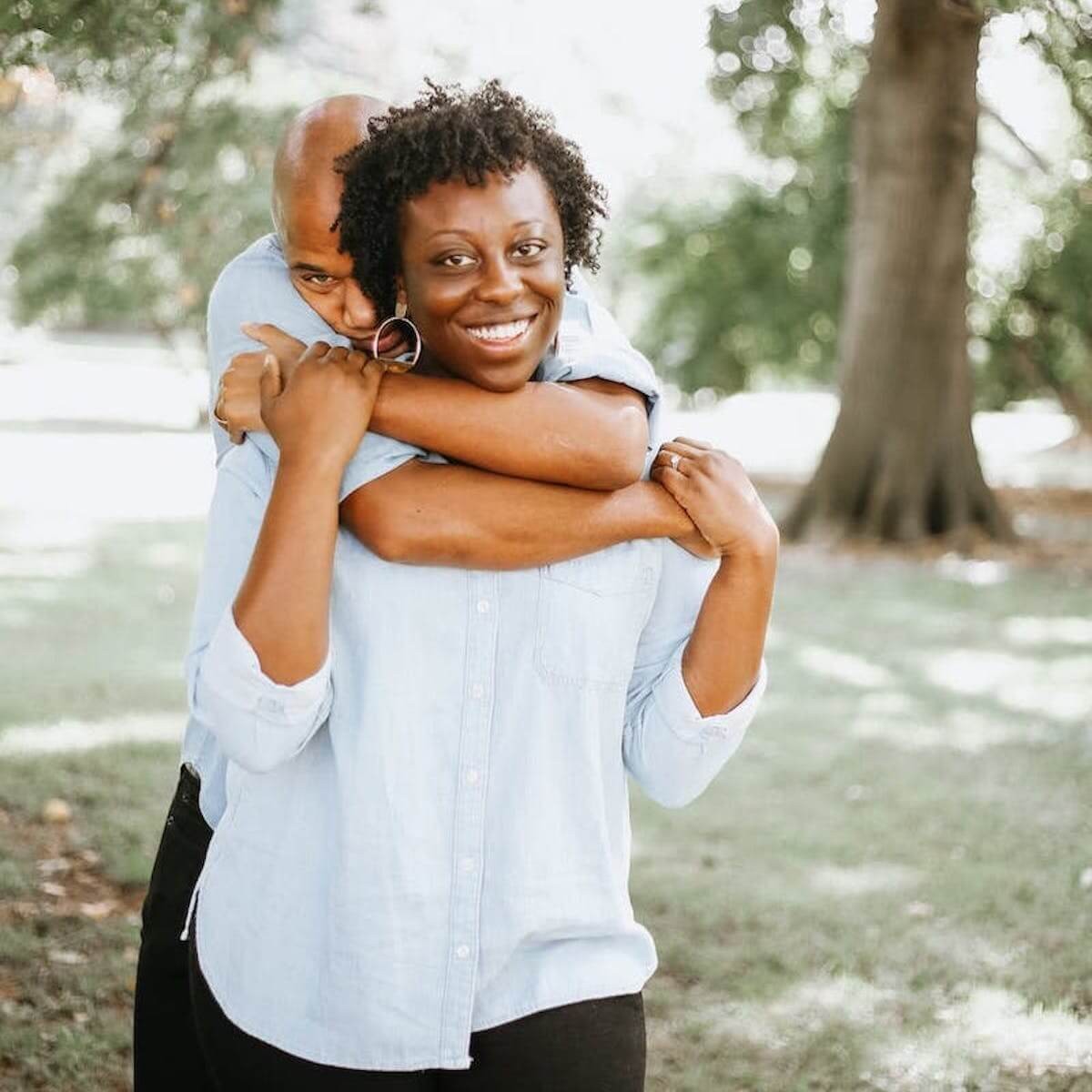 Couple smiling in park, woman in white shirt and black pants, man in blue shirt embracing her from behind