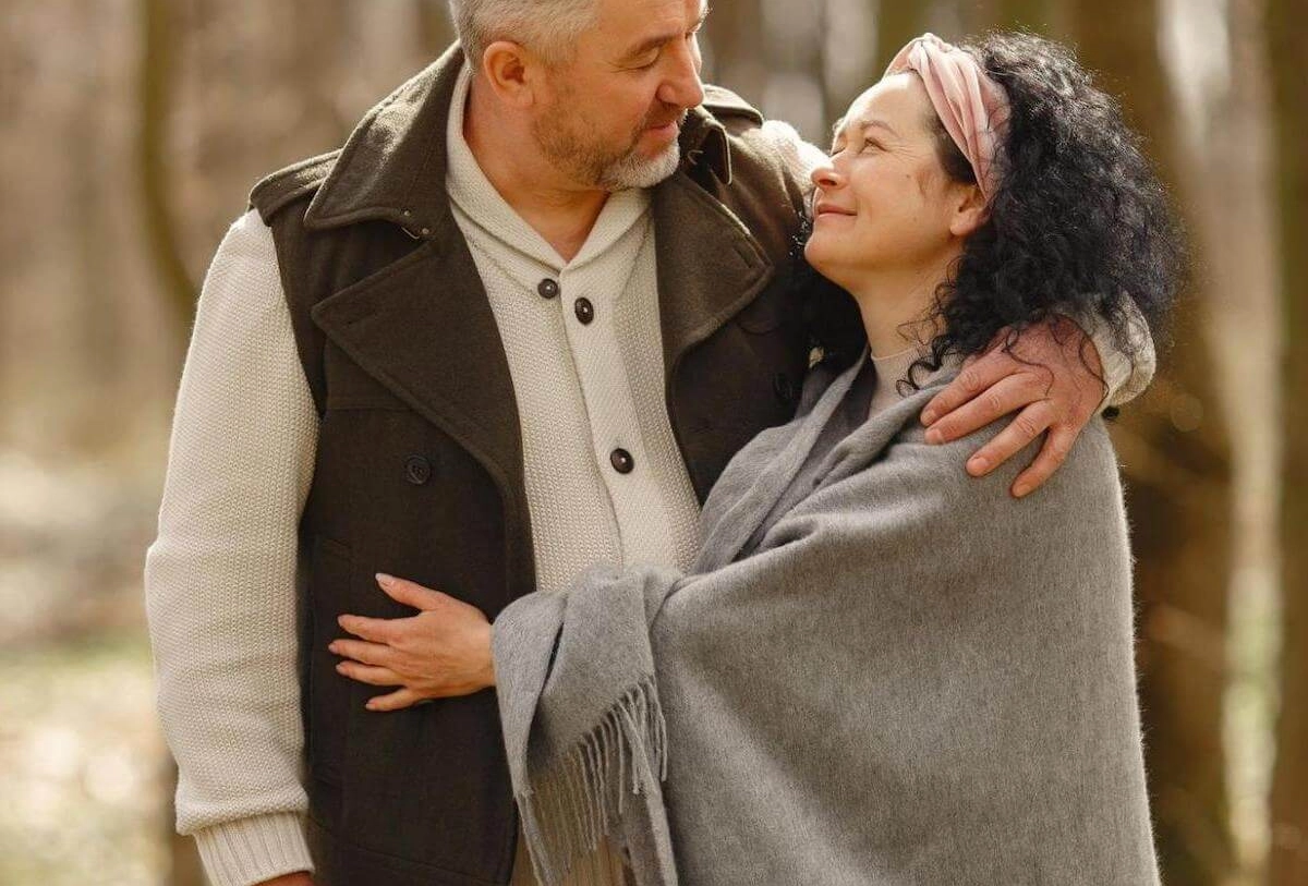 Middle-aged couple embracing outdoors, man in brown vest and cream sweater, woman in gray shawl with pink headband.