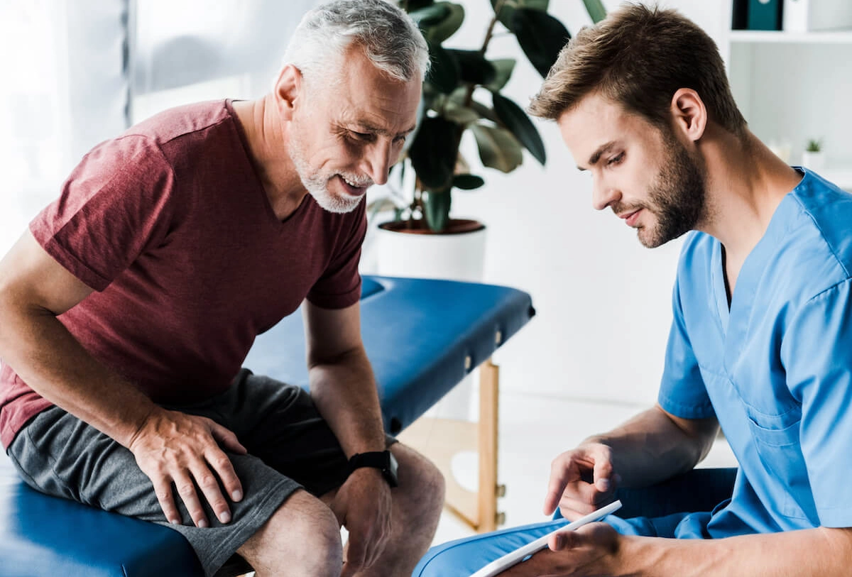 Healthcare provider in blue scrubs examining older patient's knee during medical consultation in clinic room
