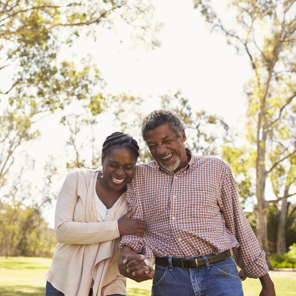 Middle-aged couple laughing together while walking through sunny park with trees