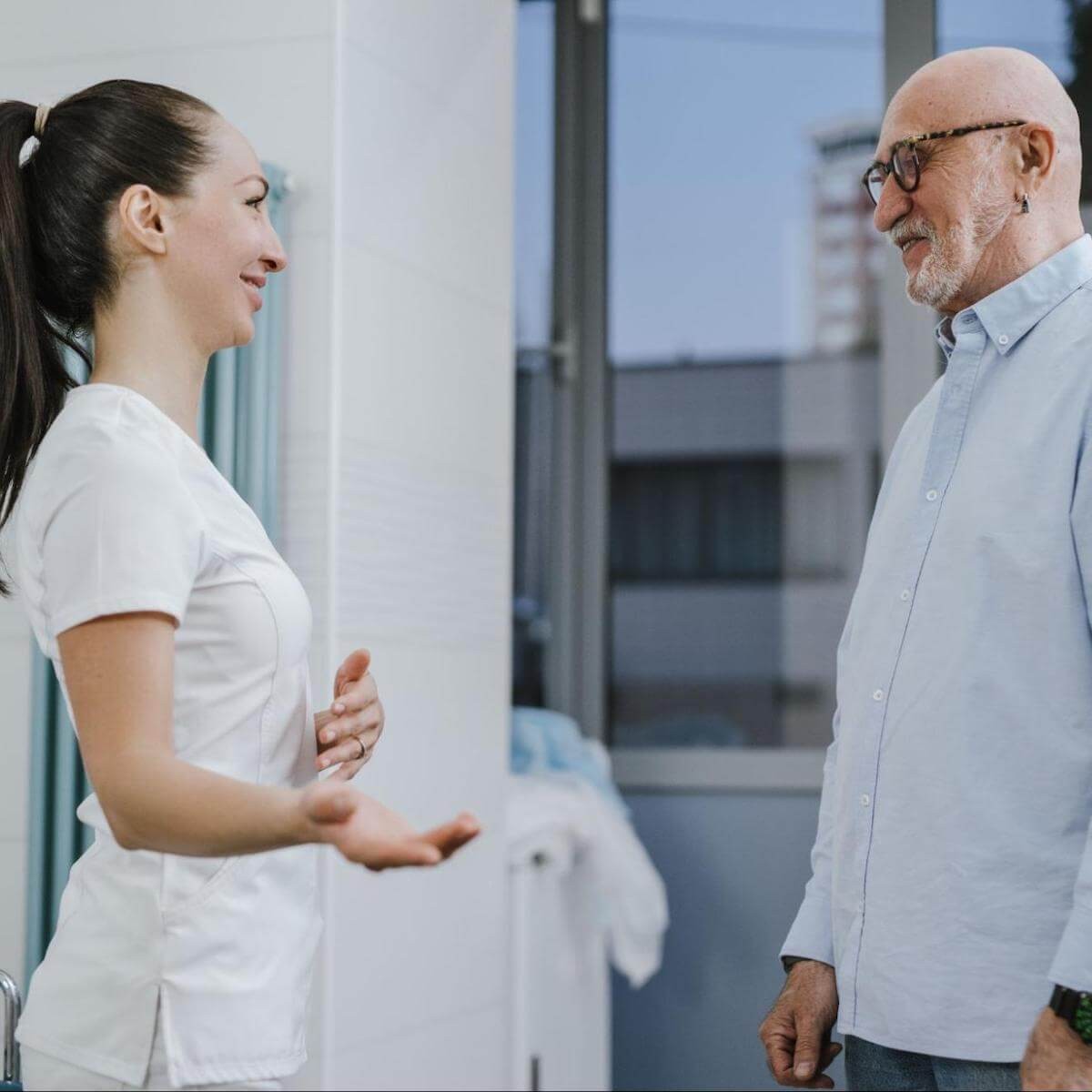 Healthcare worker in white scrubs speaking with older male patient in hospital room with city view