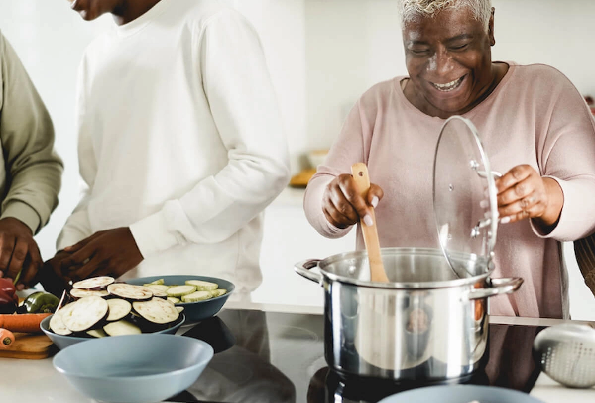Smiling woman cooking in a kitchen while someone beside her prepares sliced vegetables on the counter